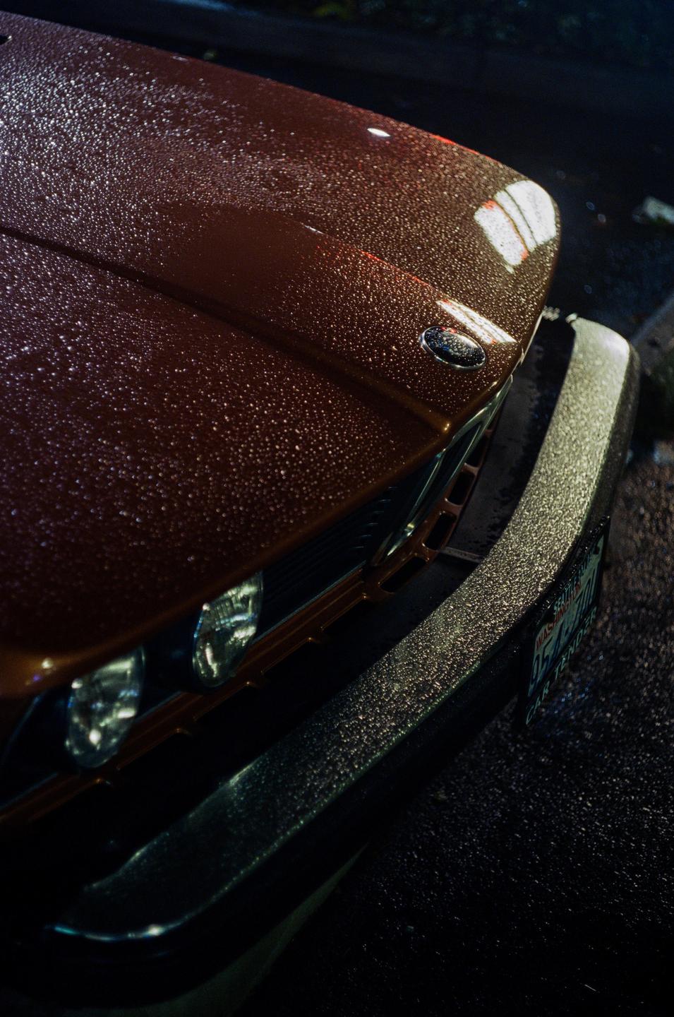Raindrops cover the hood and bumper of a brown car at night.