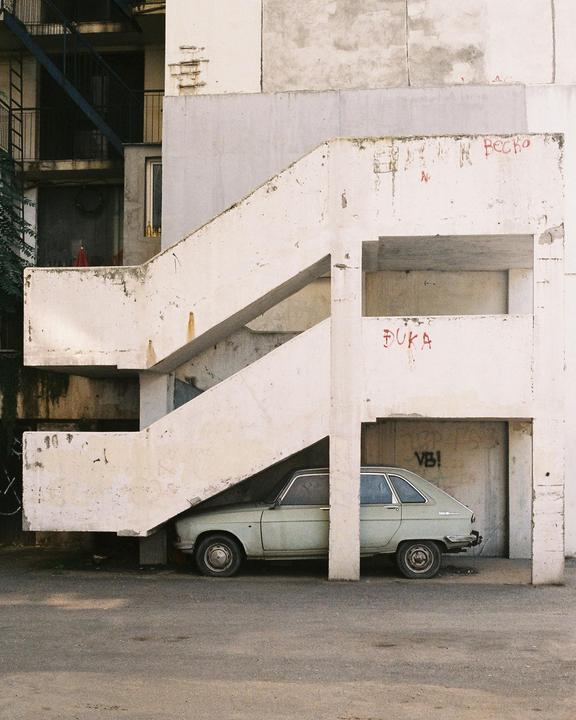 A light green car is parked under a concrete outdoor staircase with graffiti on the walls.