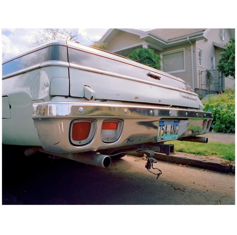 The rear of a white station wagon with a chrome bumper and an Oregon license plate is parked on a street next to a house.