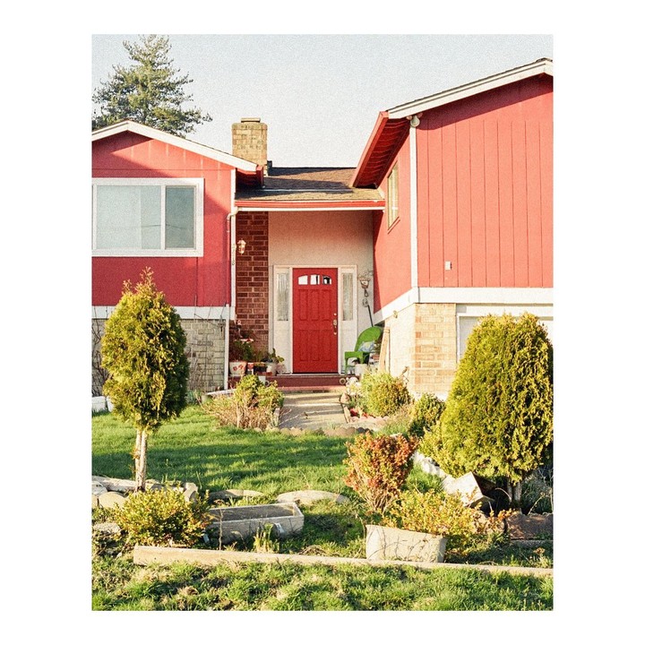 A red house with a red door is surrounded by a green lawn and shrubs under a bright sky.