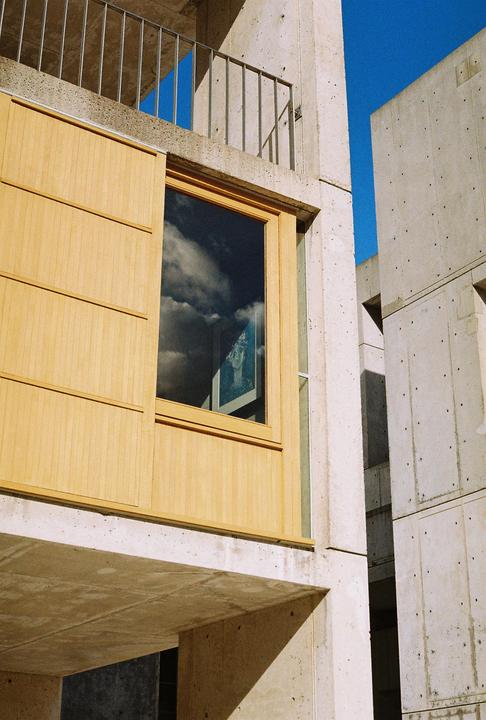 A concrete building with a wooden facade and a window reflecting clouds is shown from a low angle.