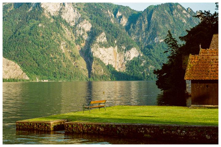 A wooden bench sits on a grassy bank next to a lake with mountains in the background.