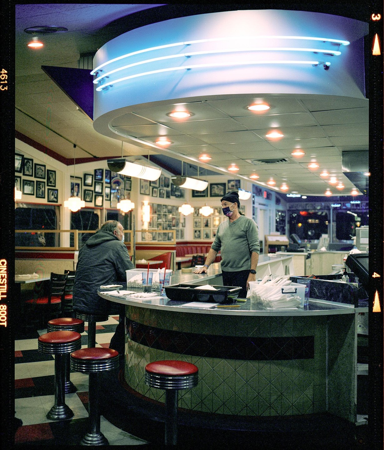 Two men are inside a diner with neon lights and red bar stools.