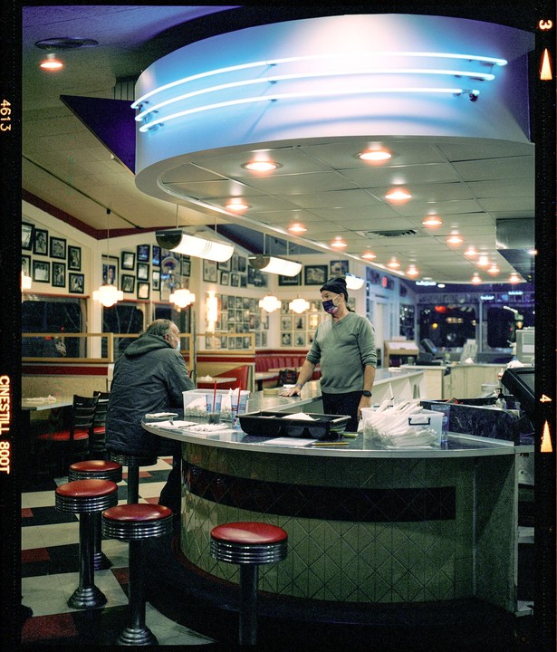Two men are inside a diner with neon lights and red bar stools.