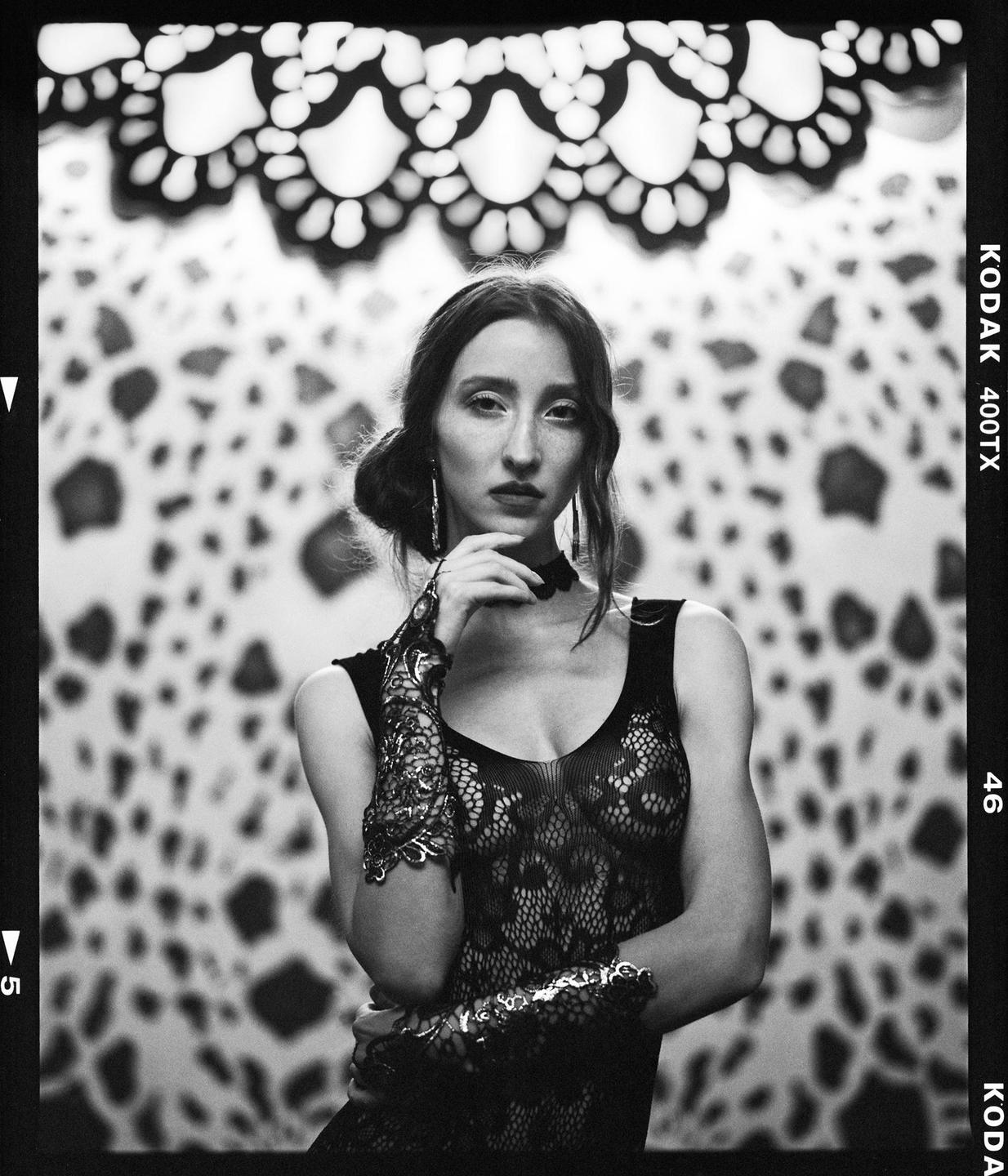 A woman in a lace top and gloves poses in front of a patterned background.