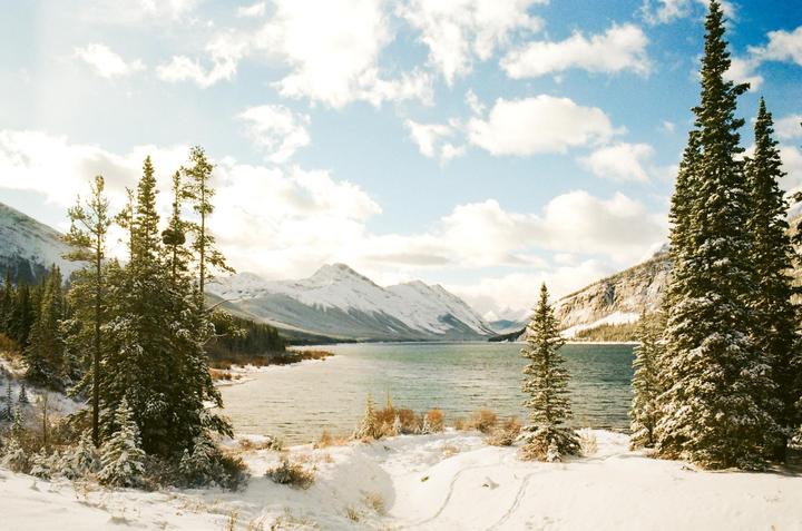A snowy landscape shows a lake surrounded by snow-covered mountains and pine trees under a cloudy sky.