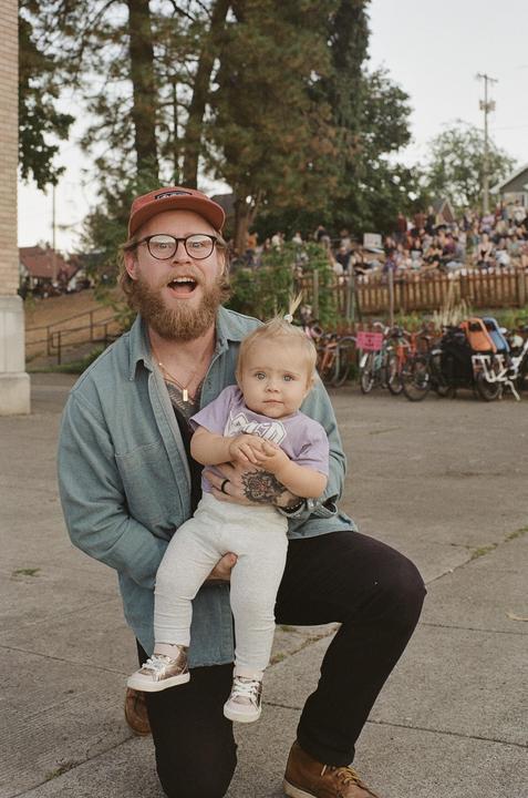 A man kneels while holding a baby with a crowd of people in the background.