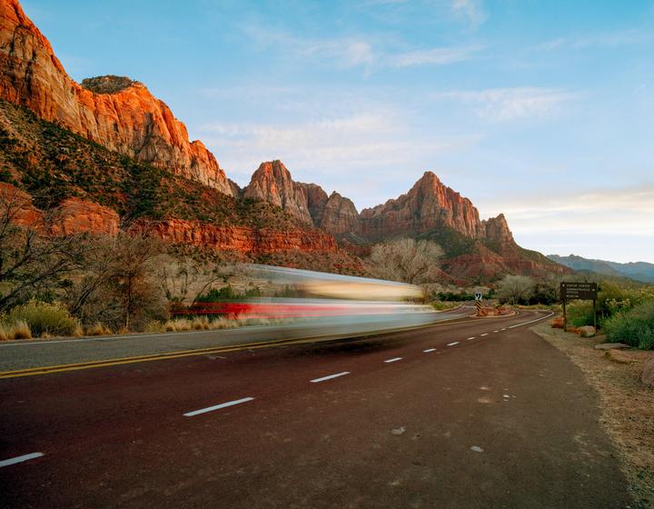 A long exposure shot of a road with light trails from a passing vehicle, set against a backdrop of red rock mountains under a blue sky.