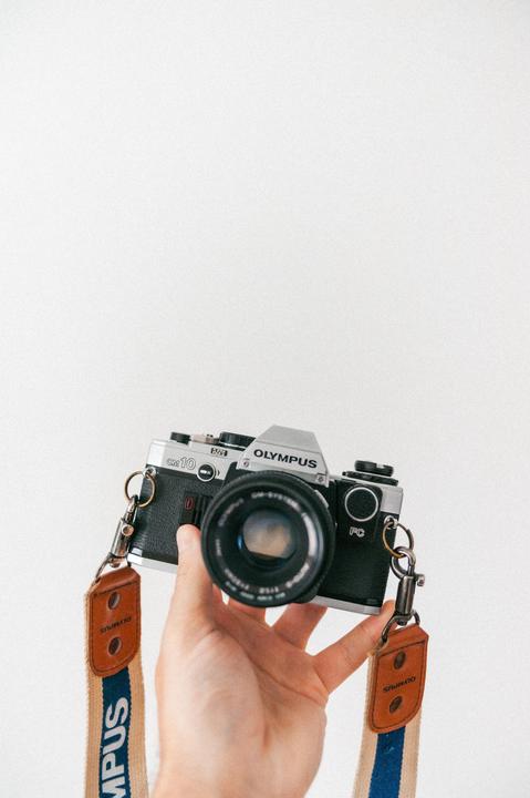 A hand holds a vintage Olympus OM10 camera with a branded strap against a white background.