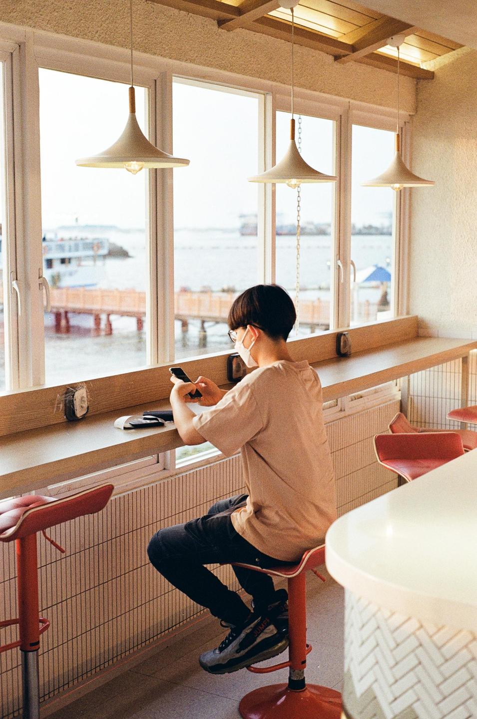 A person wearing a face mask sits at a counter by a window overlooking a body of water and uses a smartphone.