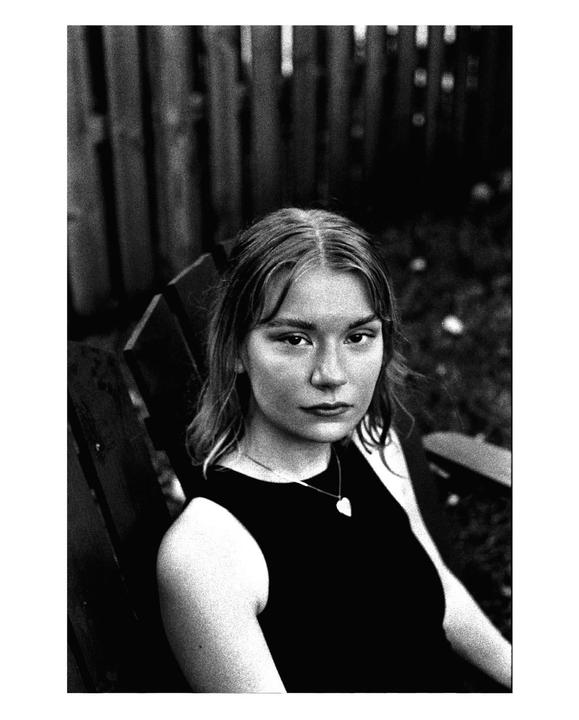 A young woman with a heart pendant necklace sits in a wooden chair outdoors.