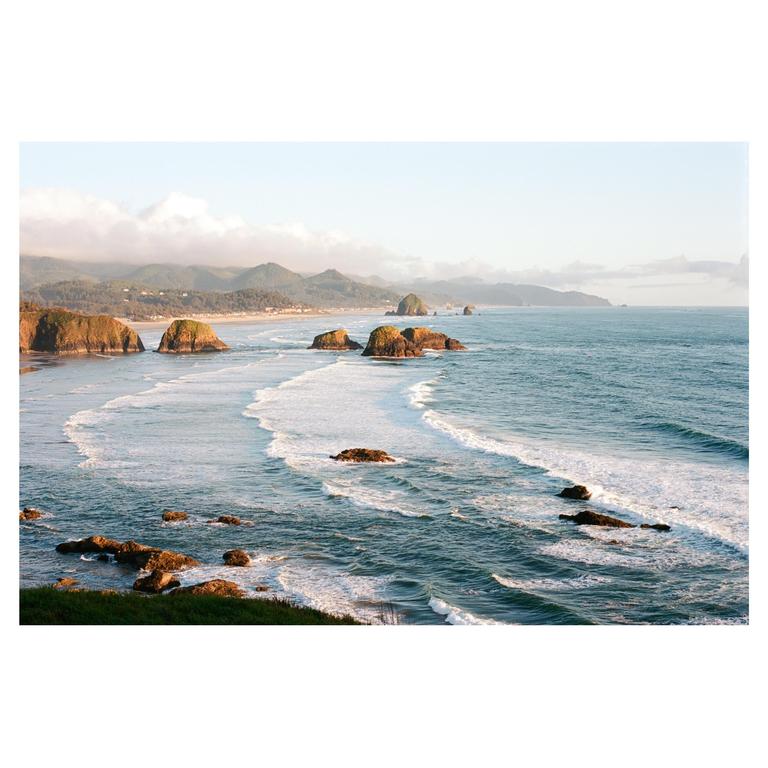 Waves crash on a rocky beach with hills and a town in the distance under a hazy sky.