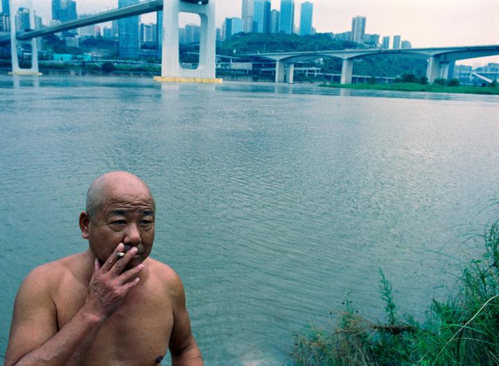 A man smokes a cigarette by a river with bridges and a city in the background.