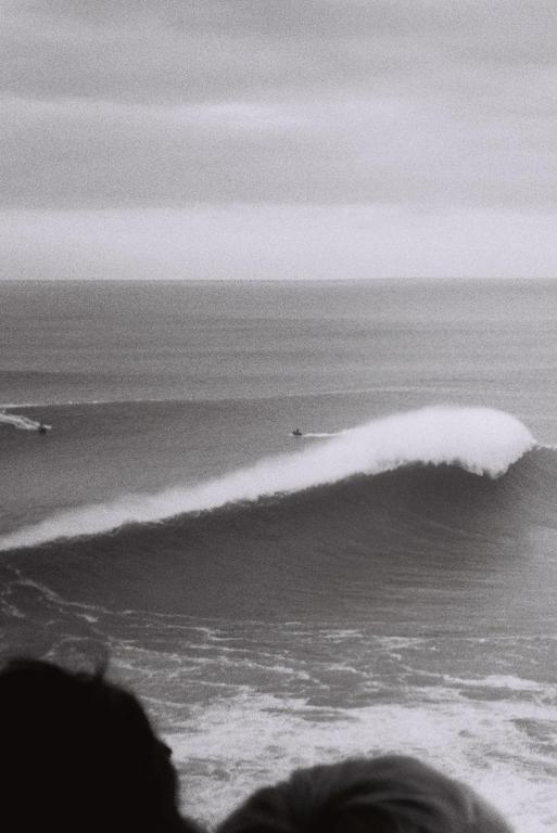 A jet ski rides a large wave in the ocean, with two heads visible in the foreground.