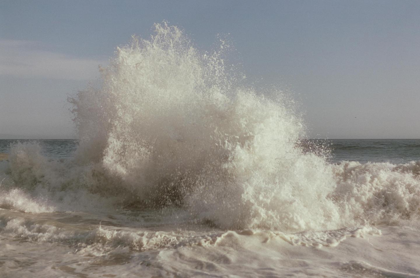 A large wave crashes on the shore, creating a burst of white spray and foam.