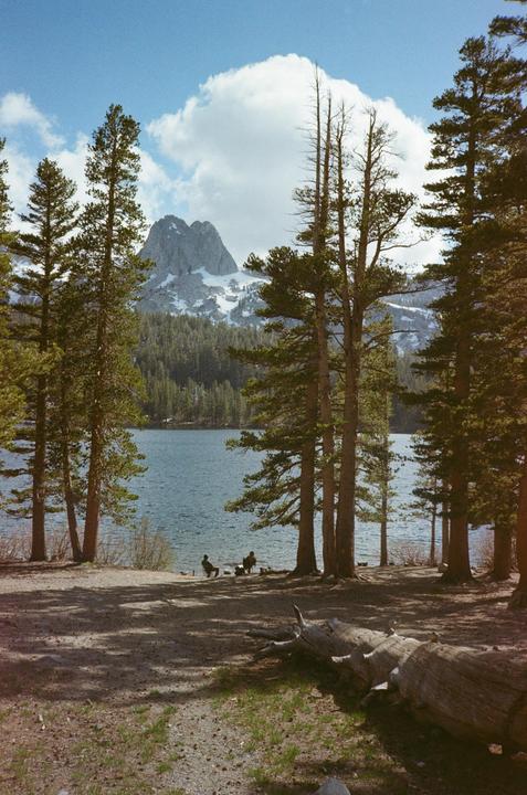 Two people sit by a lake with a snow-capped mountain and pine trees in the background.