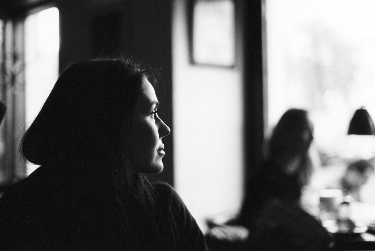 A woman is seen in profile, looking out a window, with another person out of focus in the background.