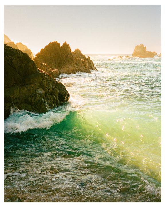 A wave crashes against a rocky coastline under a bright sky.