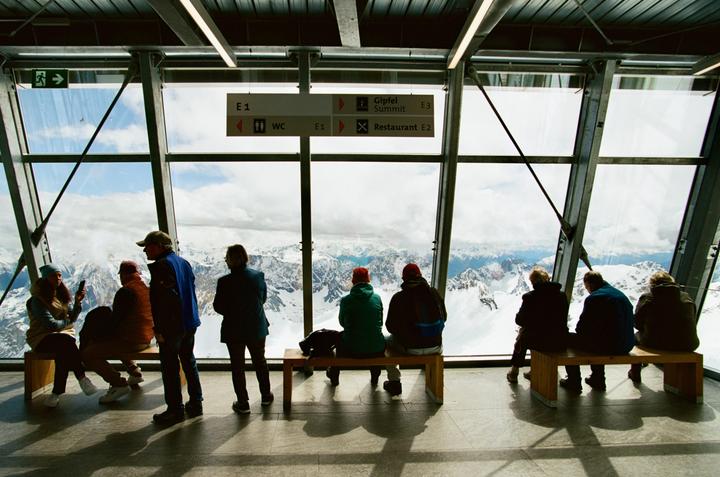 Several people are looking out of a large window at a snowy mountain range.