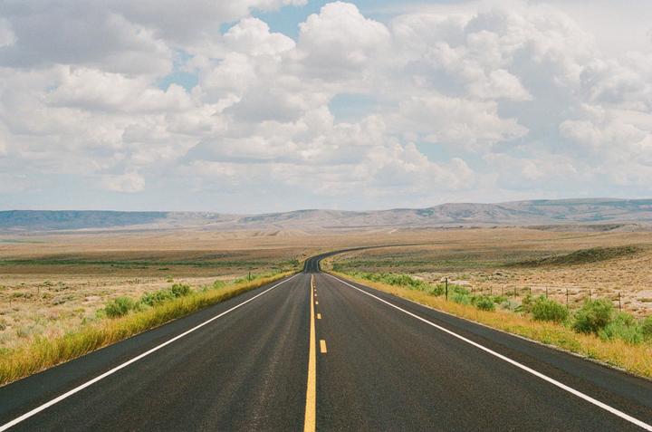 A long, empty road stretches into the distance through a vast, arid landscape under a cloudy sky.