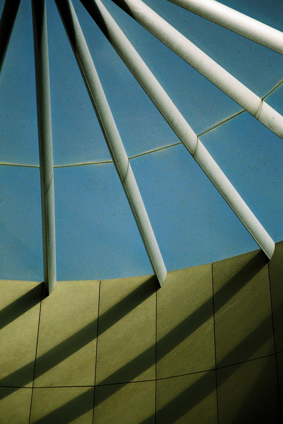 Sunlight casts shadows from white beams onto a curved wall under a blue sky.