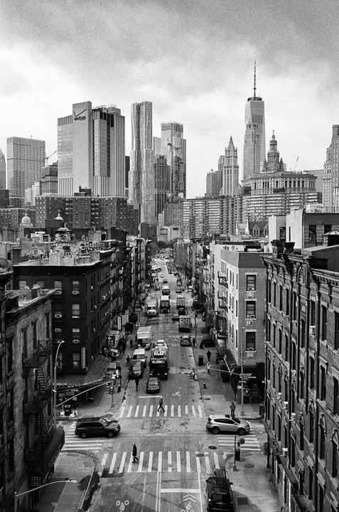 A high-angle view shows a busy street with cars and pedestrians, flanked by buildings, with a city skyline in the background.