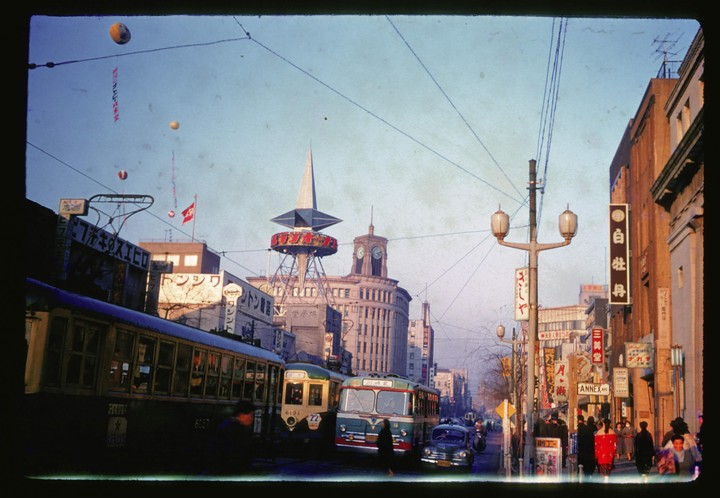 A busy street in Tokyo is filled with trams, buses, cars, and pedestrians, with a prominent clock tower and a spire-topped building in the background.