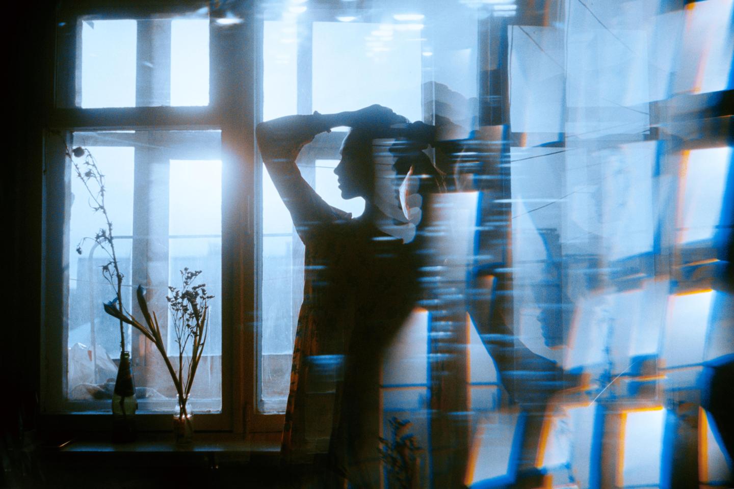 A woman stands in silhouette in front of a bright window with dried plants on the sill, with a blue and orange light leak effect.