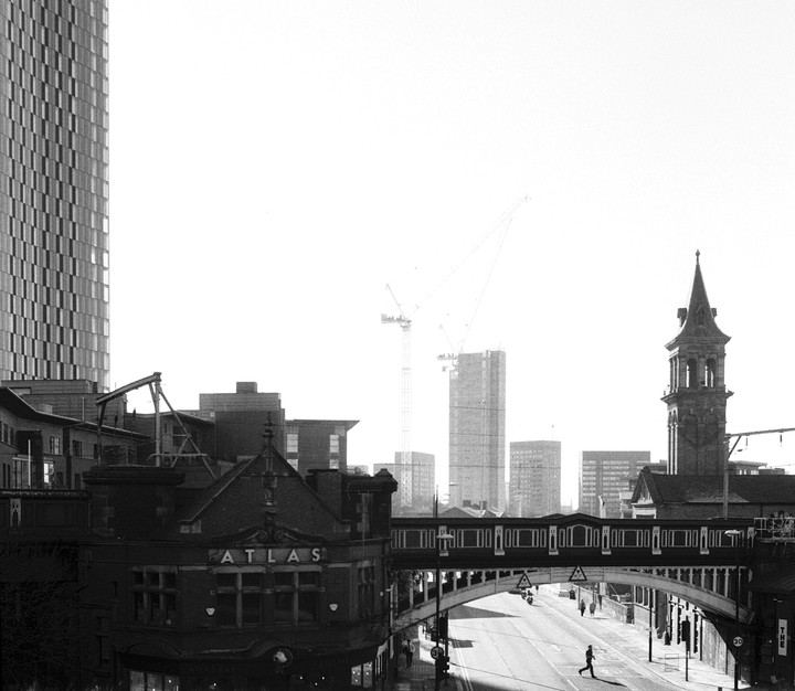 A street scene in a city shows a pedestrian bridge over a road, with a tall building and a church tower visible.