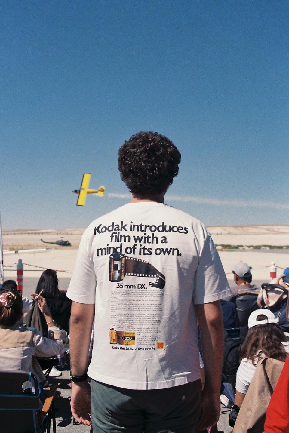 A man wearing a white t-shirt with a Kodak advertisement on the back watches a yellow airplane fly across a clear blue sky.