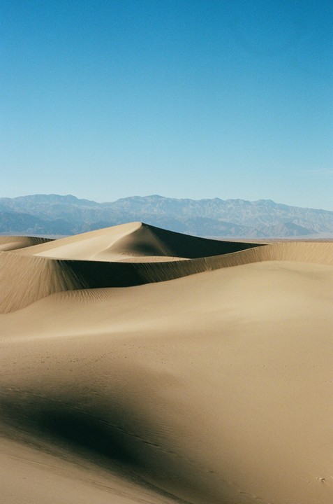 A vast desert landscape features sand dunes with sharp shadows and distant mountains under a clear sky.
