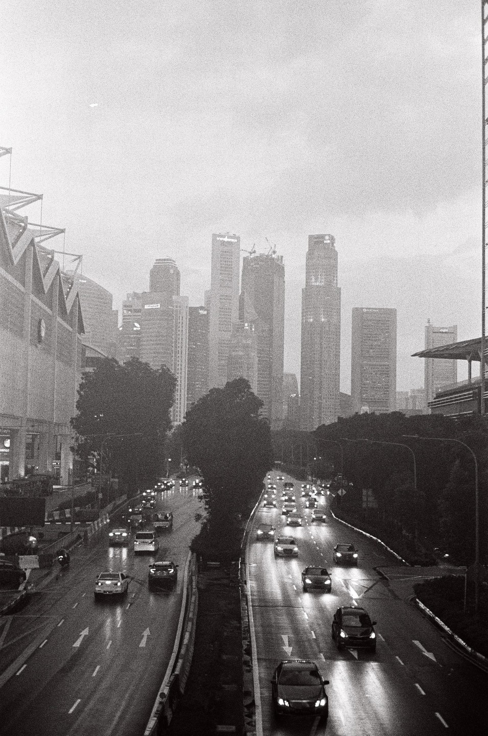 A busy highway with cars driving in both directions is wet from rain, with a city skyline in the background under a hazy sky.