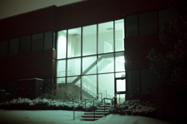 A large glass window of a brick building reveals an interior staircase, with snow-covered ground and bushes in the foreground.