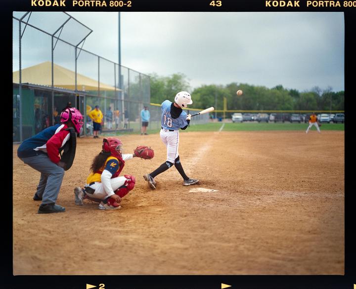 A baseball player swings a bat at a ball while a catcher and umpire watch on a dirt field.
