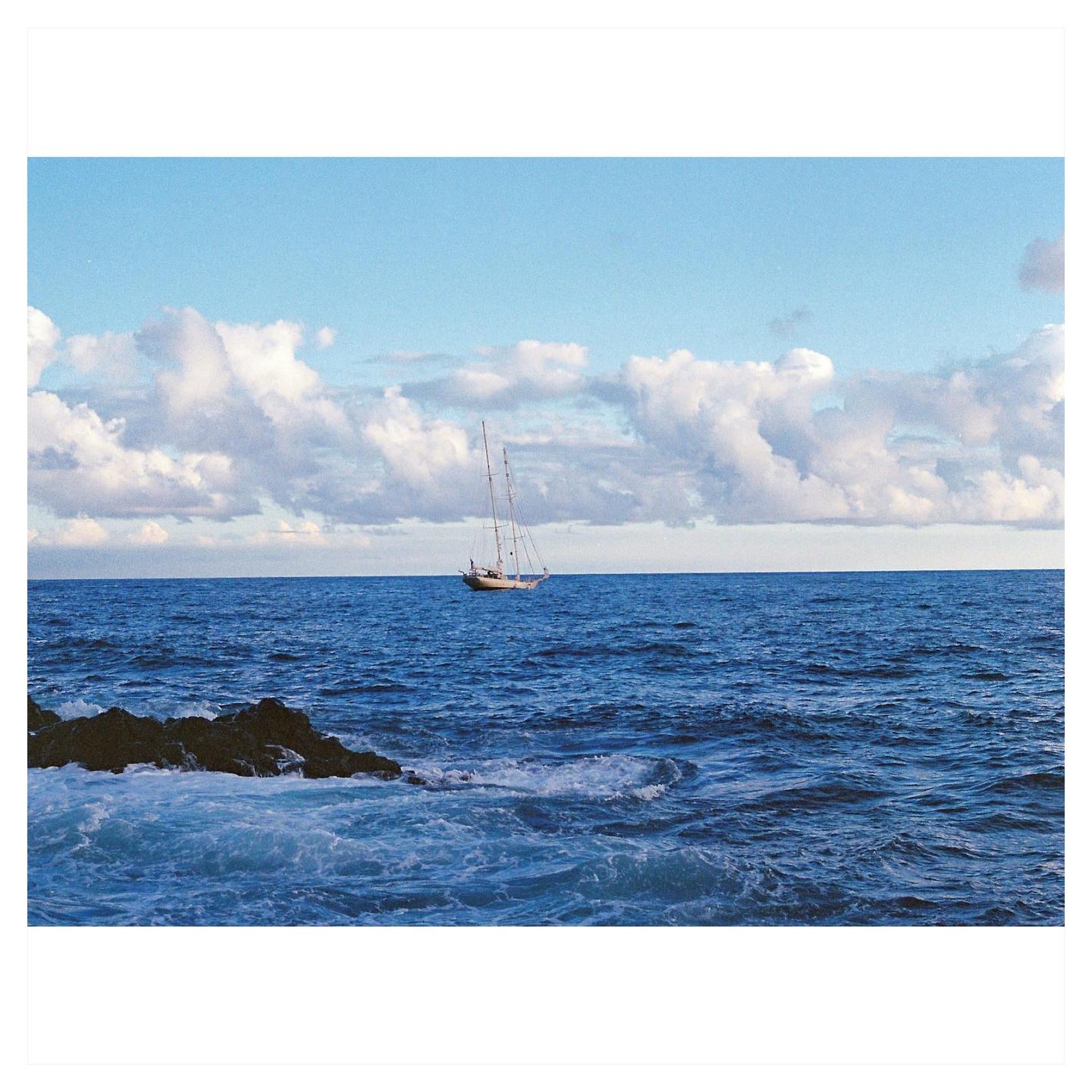 A sailboat floats on the ocean with a rocky shore in the foreground under a cloudy sky.