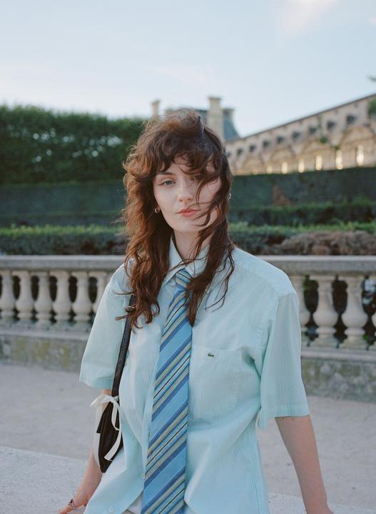 A woman with long, curly hair and a striped tie poses outdoors with a balustrade and building in the background.