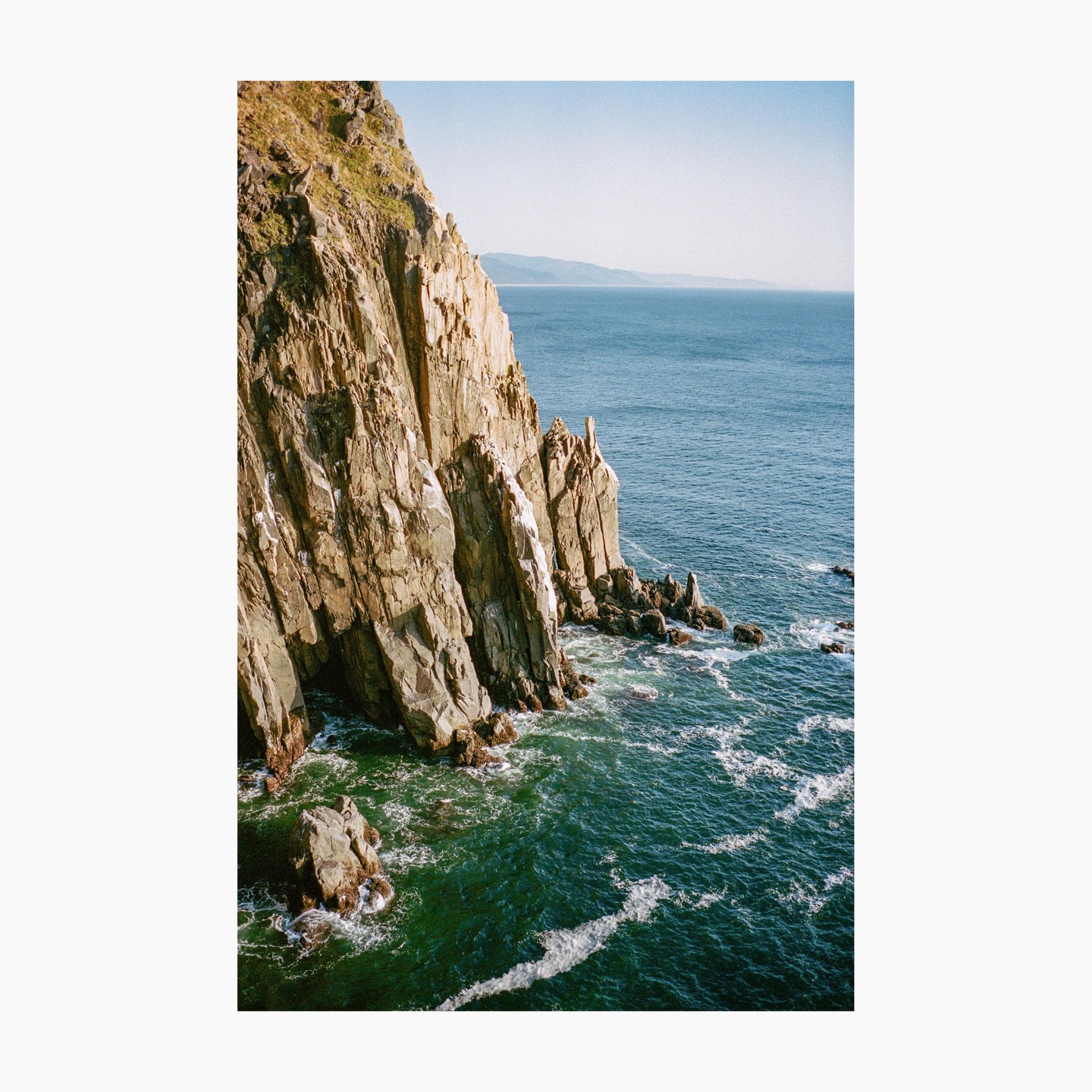 A steep, rocky cliff face meets the ocean, with waves crashing against the shore and a distant coastline visible under a clear sky.