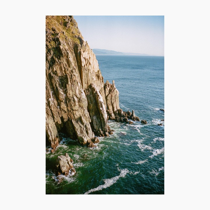 A steep, rocky cliff face meets the ocean, with waves crashing against the shore and a distant coastline visible under a clear sky.