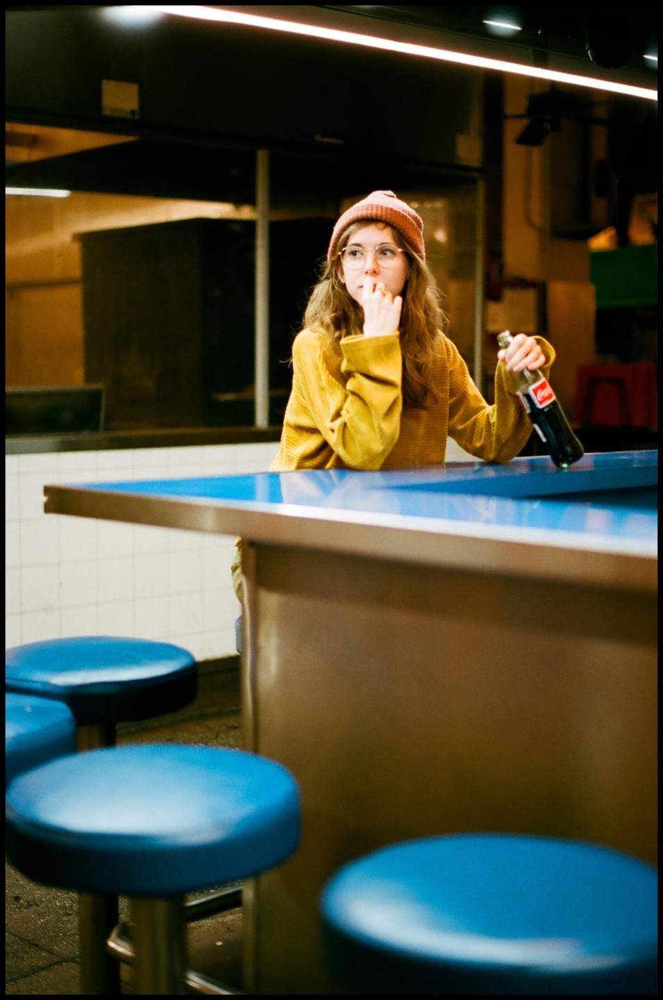 A woman in a beanie and glasses sits at a diner counter with a bottle of soda.