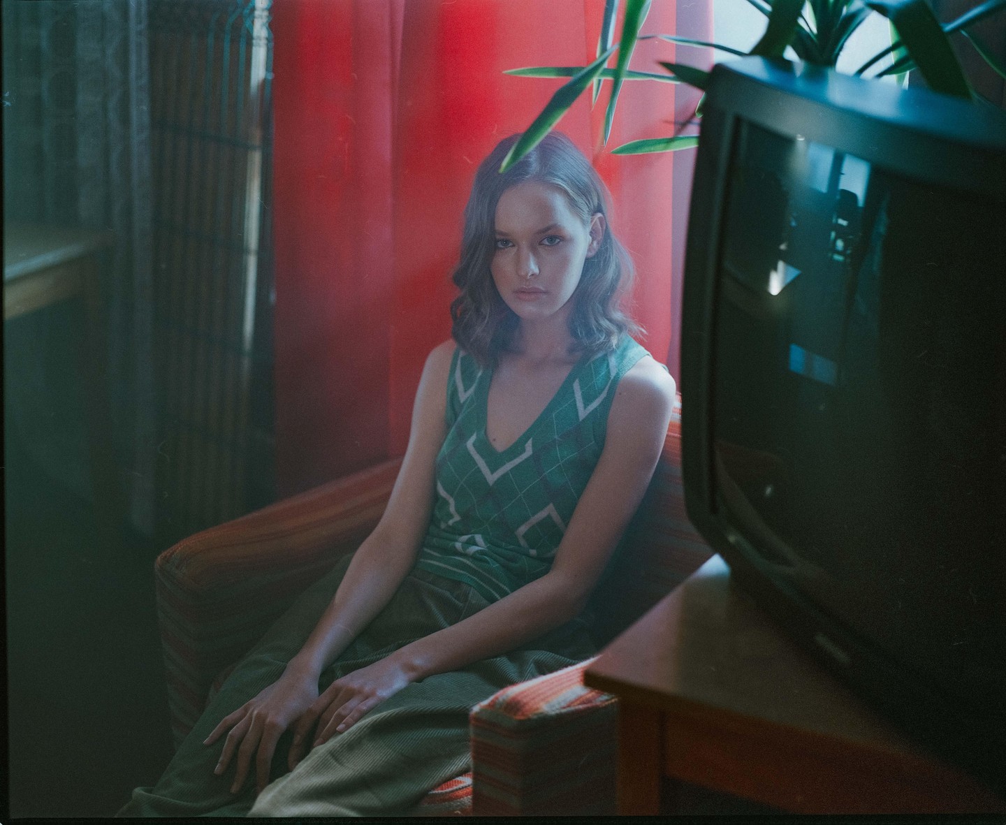 A woman with dark hair sits in an armchair next to a television set and a plant.