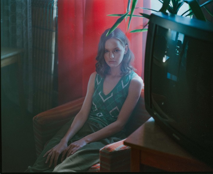 A woman with dark hair sits in an armchair next to a television set and a plant.