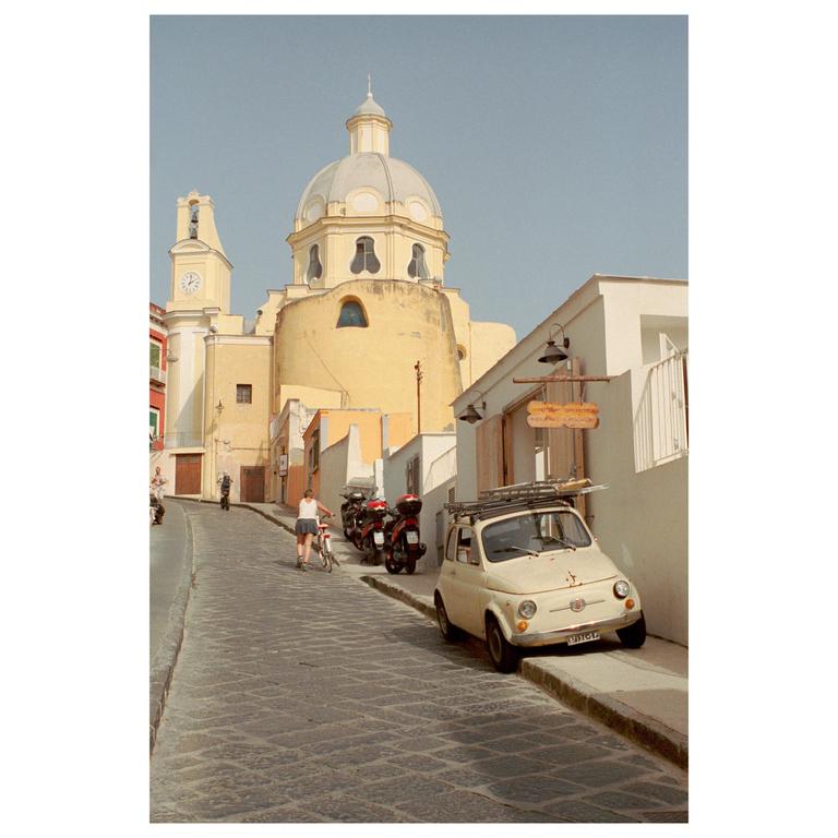A woman pushes a bicycle up a cobblestone street with a small car parked on the right and a large domed building in the background.
