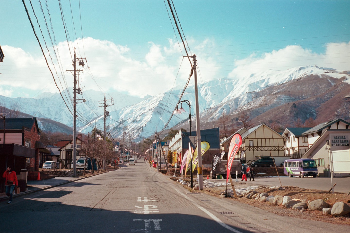 A street with buildings and power lines leads toward snow-covered mountains under a blue sky with clouds.