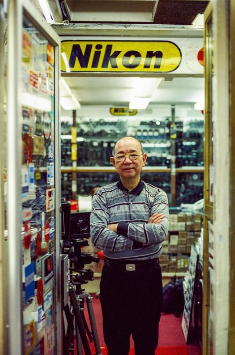 A man with glasses and a striped shirt stands with his arms crossed in a camera store.