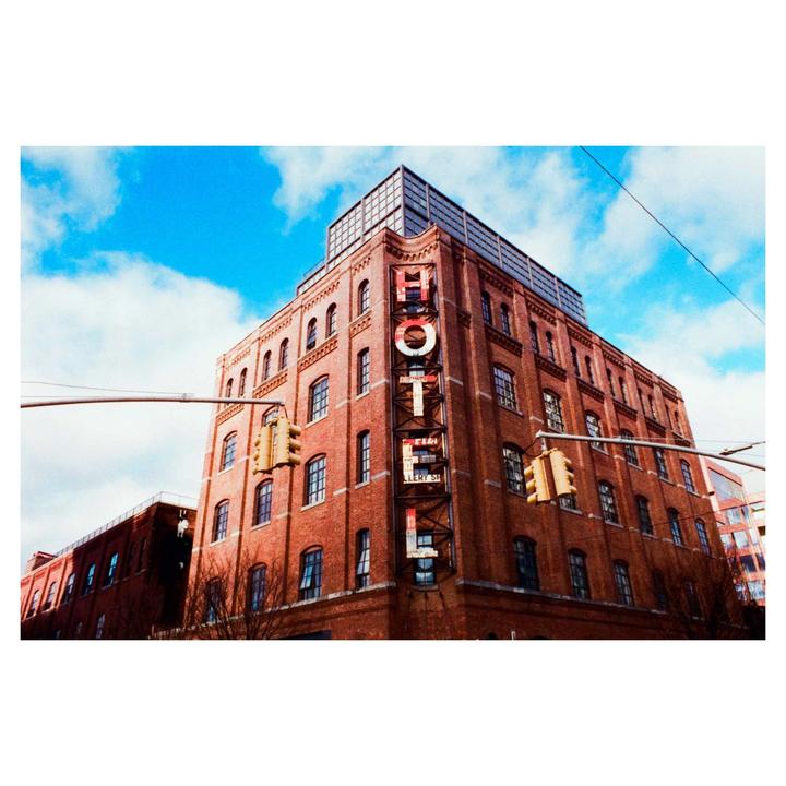 A brick building with a neon "HOTEL" sign on its side stands against a blue sky with white clouds.