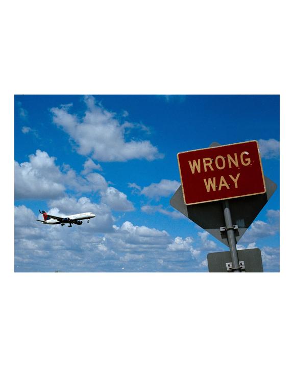 A red wrong way sign is in the foreground with an airplane flying in a blue sky with clouds in the background.