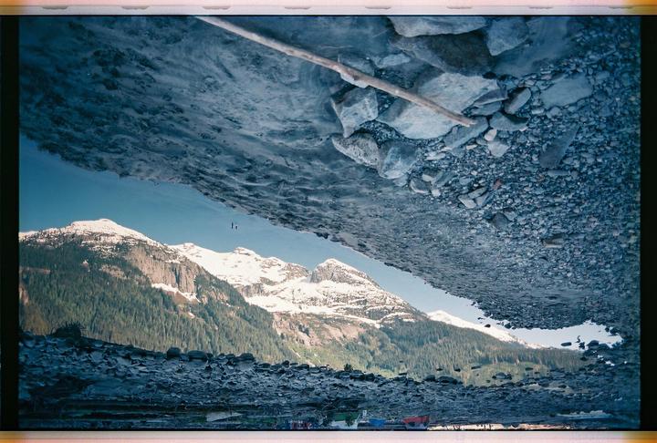 A reflection of snow-capped mountains and a forest is visible in a body of water, surrounded by rocks and pebbles.