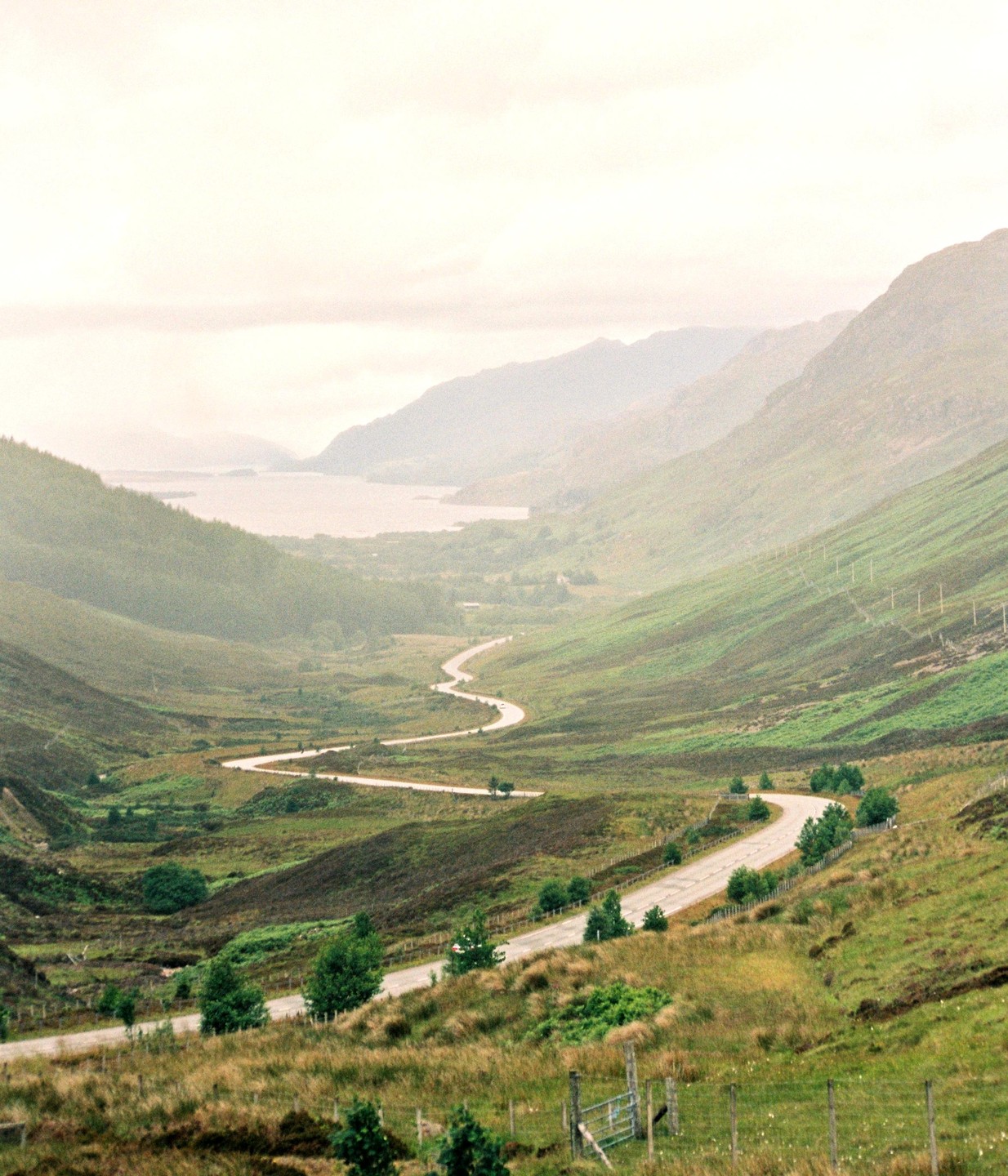 A winding road descends through a valley towards a body of water surrounded by mountains.