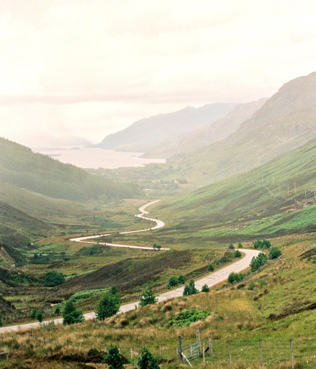 A winding road descends through a valley towards a body of water surrounded by mountains.