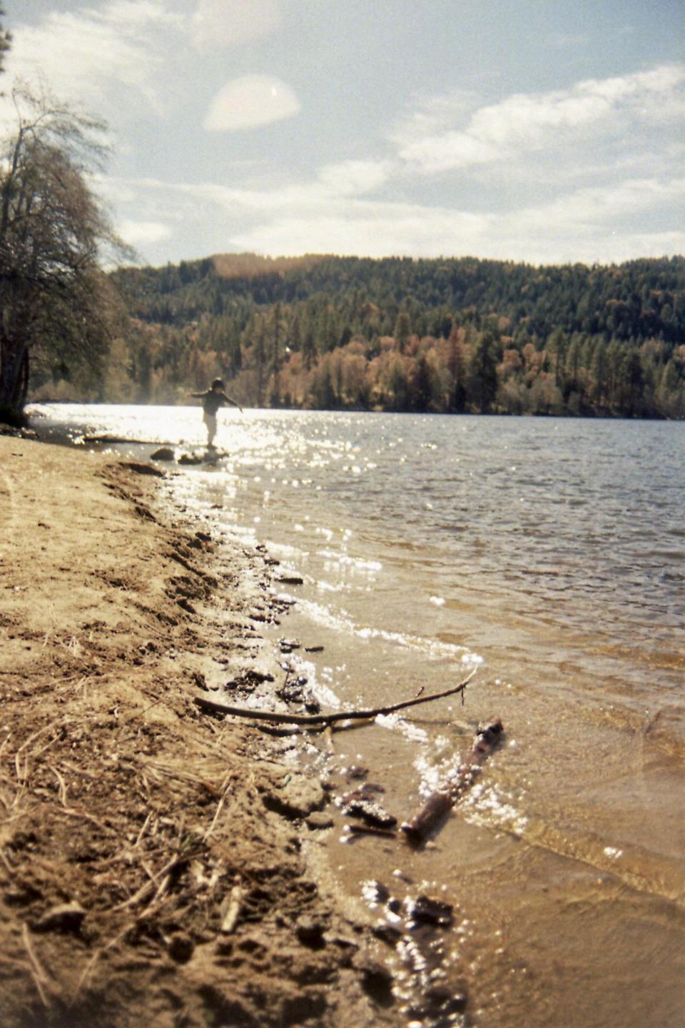 A child stands at the edge of a lake with arms outstretched, with a forest on the far bank.